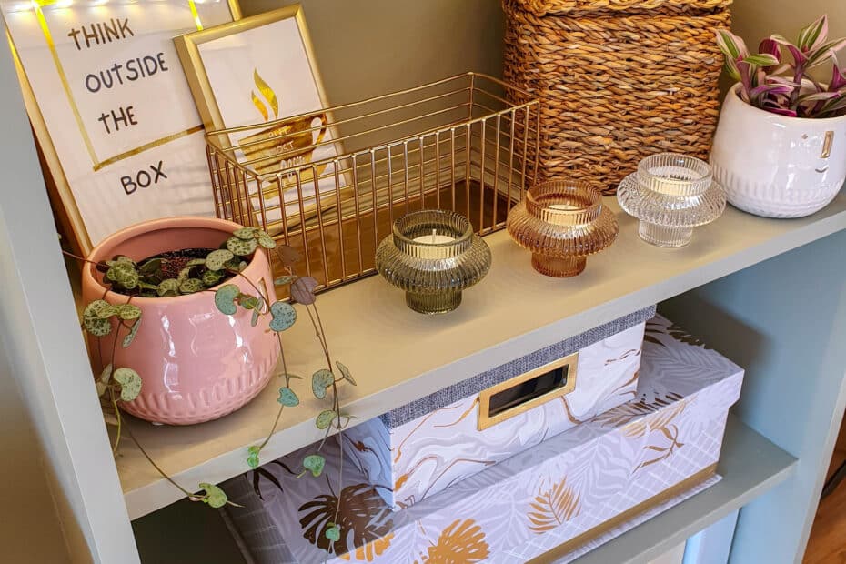 A shelf displays a framed “Think Outside the Box” sign—a reminder to avoid the 5 Mistakes Marketers Make with Pinterest—plus a golden wire basket, potted plants, decorative candle holders, a woven basket, and colourful binders on the bottom shelf.