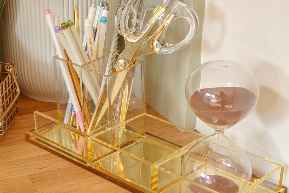 A gold and clear acrylic organiser holds coloured pens, pencils, scissors, and a ruler on a wooden desk—perfect for jotting down insights like the 5 Mistakes Marketers Make with Pinterest. An hourglass and white plant pot complete the tidy scene.