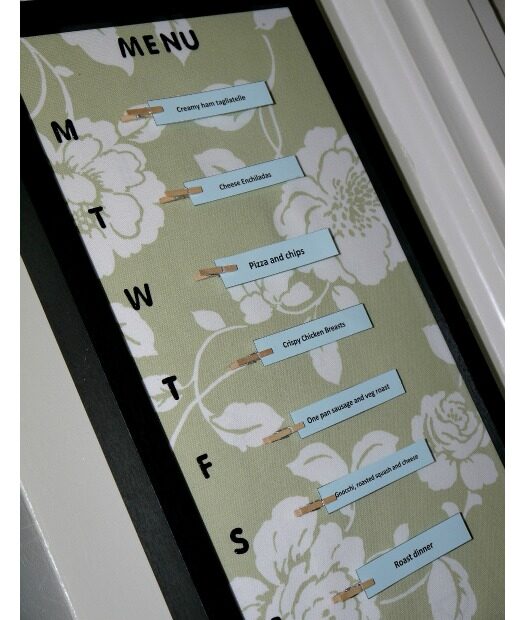 A framed weekly menu board with floral background shows meal plans for each day. Small cards clipped under initials M to S list dishes like pizza and pasta—perfect inspiration if you’re wondering how to make a meal plan board yourself.