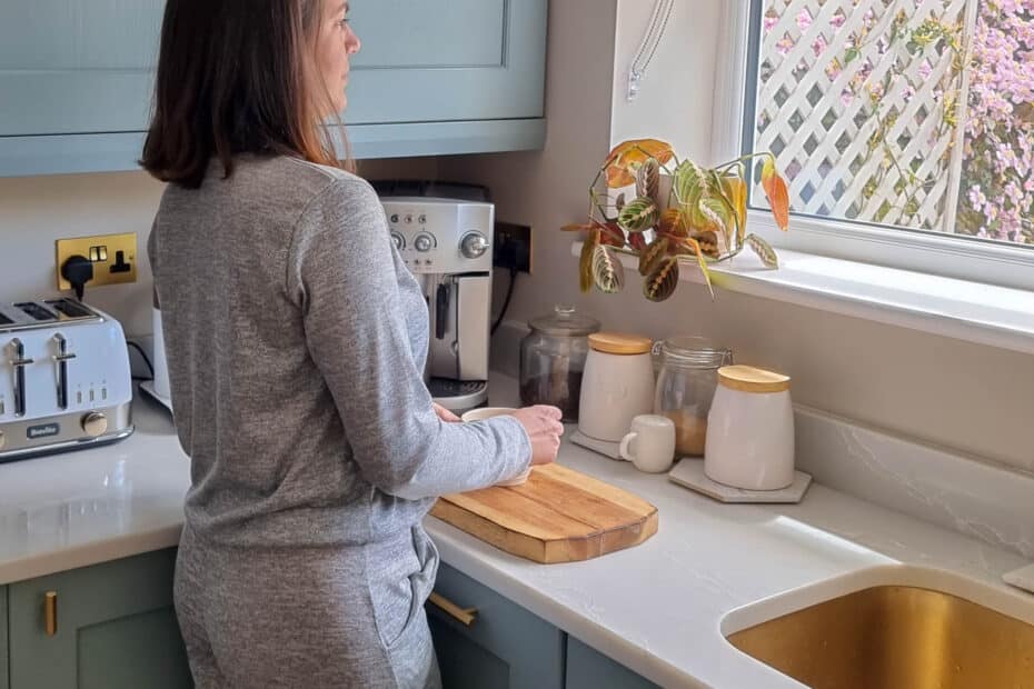 A woman in grey loungewear stands in a kitchen by a worktop, facing a window with sunlight streaming in. There is a chopping board, a coffee machine, a toaster, jars, and a potted plant on the worktop.