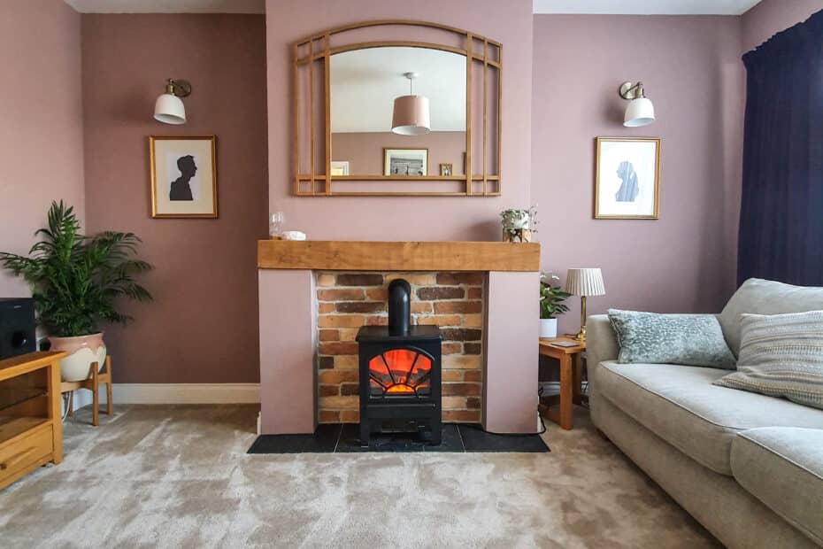 A cosy living room perfect for staging the inside of your home, featuring a beige carpet, mauve walls, a brick fireplace with a wood-burning stove, wooden mantelpiece with mirror above, framed art on each side, grey sofa, plants, and soft lighting.