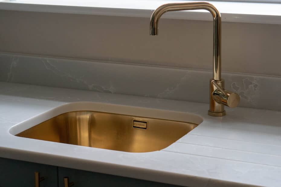A modern kitchen sink with a gold-coloured tap and basin, set into a white marble worktop with matching splashback—an elegant upgrade that can help you save money when moving by adding value to your home.