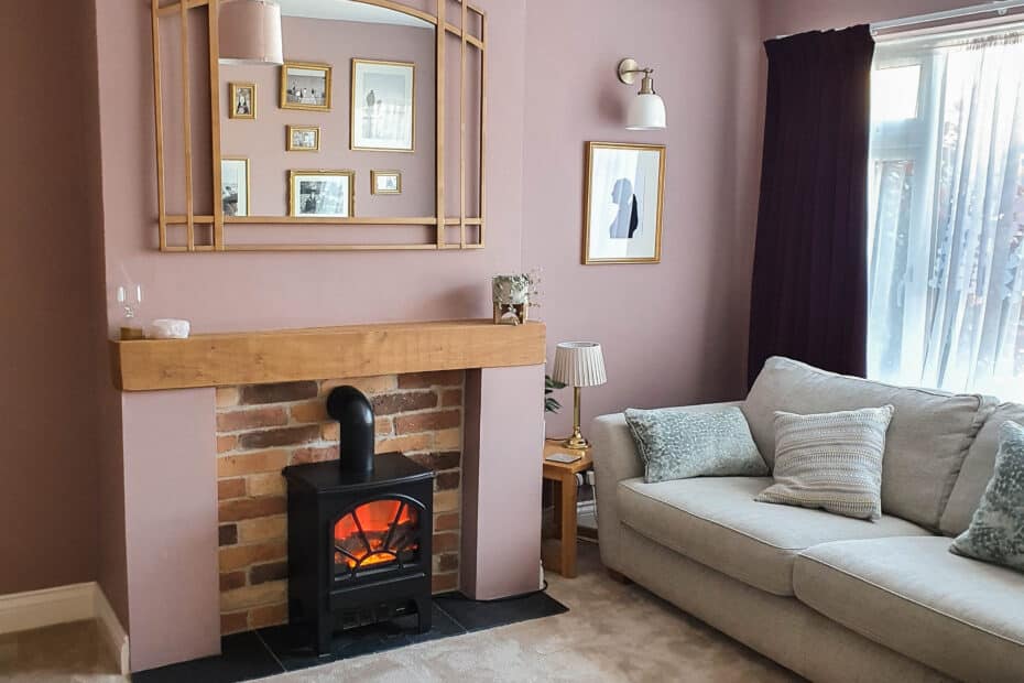 Give your room a fresh look with this cosy living room featuring a beige carpet, light sofas with cushions, a brick fireplace with a wooden mantelpiece, an electric stove heater, framed pictures, and a large mirror above the mantelpiece. Natural light fills the space.