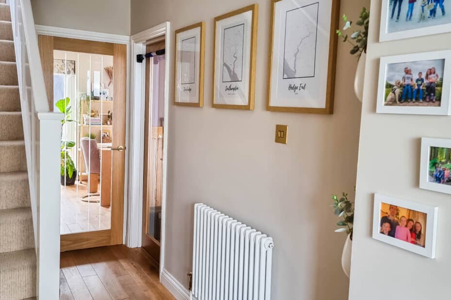 A hallway with wooden floors, a carpeted staircase, framed maps and family photos on cream walls, a white radiator, and a view into a bright, plant-filled room beyond a glass door—perfect for preparing your home to be listed.