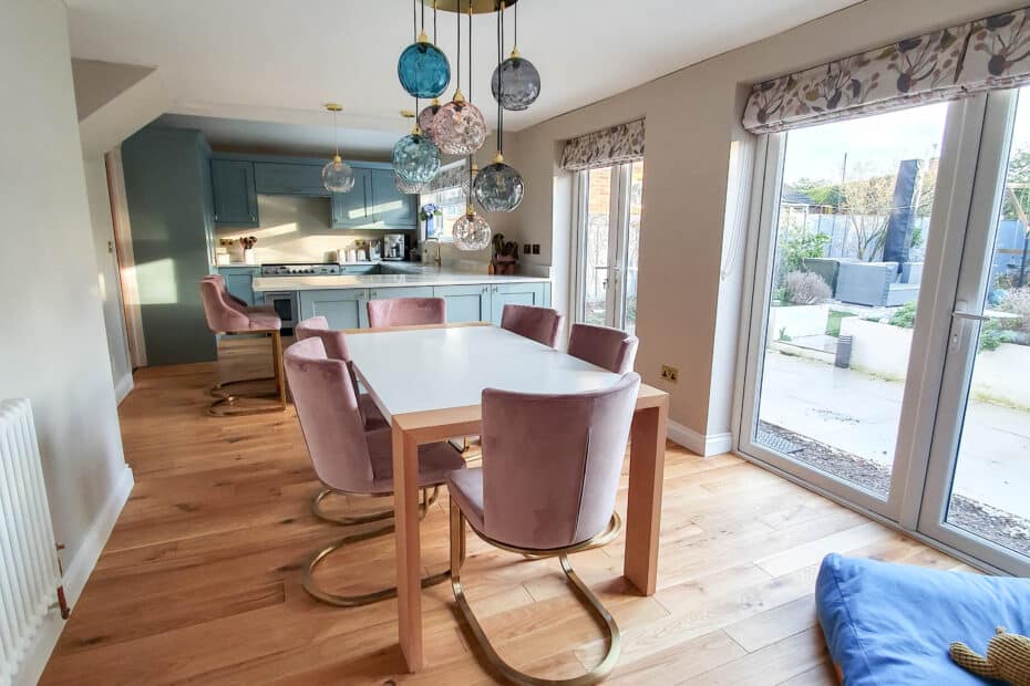 Modern dining room with light wood flooring, a white rectangular table, pink velvet chairs with gold legs, and decorative glass pendant lights. Large windows and a blue dog bed create a sense of a bigger kitchen without moving house.