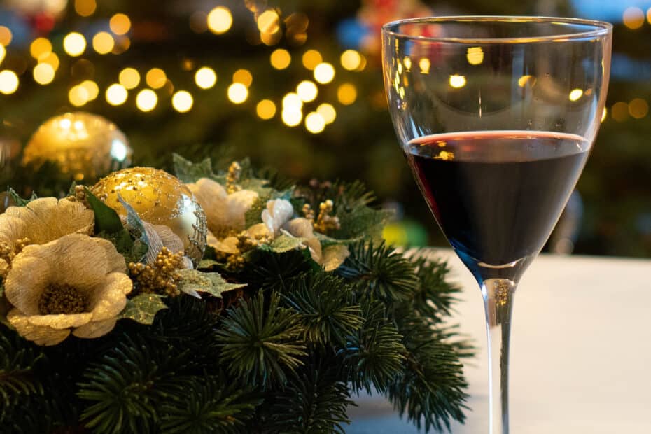 A glass of red wine sits on a table beside a festive pine and gold floral arrangement, the perfect touch when hosting Christmas Dinner, with blurred Christmas lights on a decorated tree glowing warmly in the background.