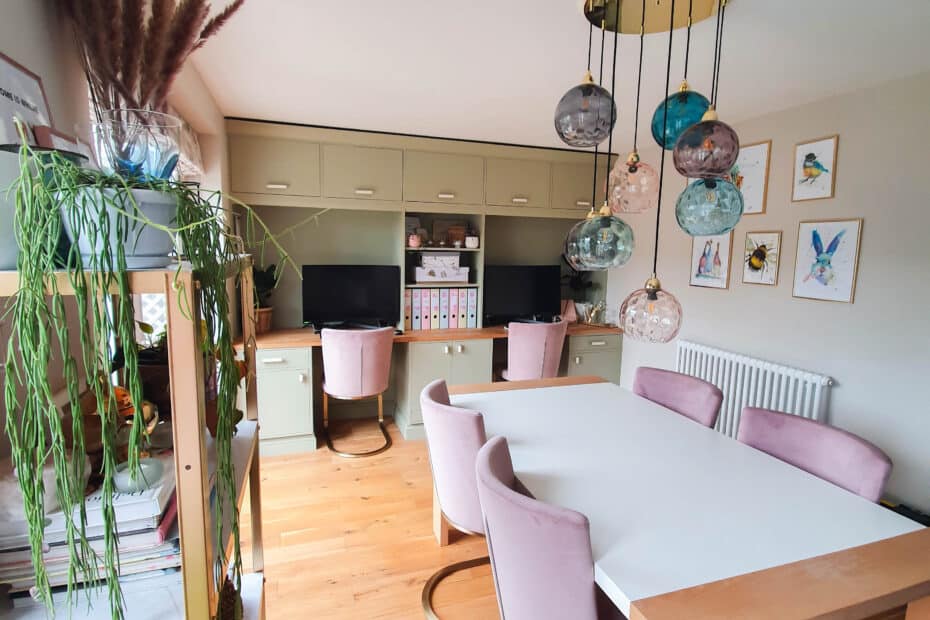 Modern home office with green cabinetry, two desks with computers, a white table surrounded by pink chairs, hanging globe lights, plants, and framed art on beige walls. Wooden floor and organised shelves offer your home an easy upgrade.