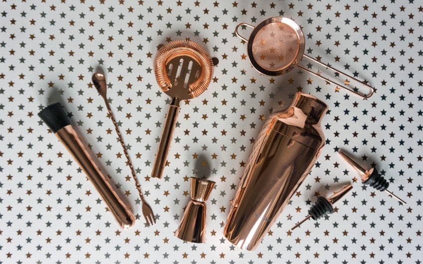 A set of copper cocktail tools for your home bar, including a shaker, strainer, jigger, muddler, stirring spoon, and pourers, arranged on a white surface with a gold star pattern.