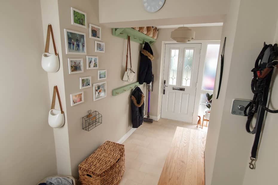 A tidy hallway perfect for buying a family home, featuring beige walls, wall art, hanging mugs, a coat rack with coats, a wicker basket, a bench, and a white front door with frosted glass panels and a clock above it.
