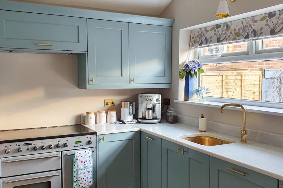 A modern kitchen with blue cupboards, a silver cooker, and a gold tap—perfect for Success With A Holiday Let. Sunlight streams through a window with a floral blind, while a vase of purple flowers adds charm to the inviting space.
