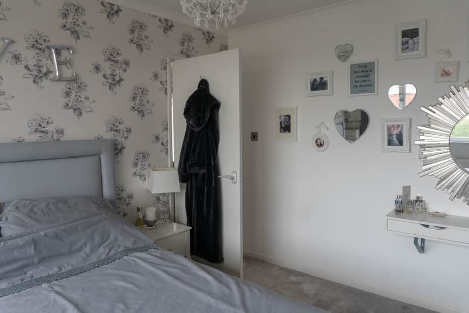 A cosy guest area in your home with a grey bed, floral wallpaper, a lamp, and framed photos on white walls. A black dressing gown hangs on the open door, with a decorative mirror and dressing table to the right.