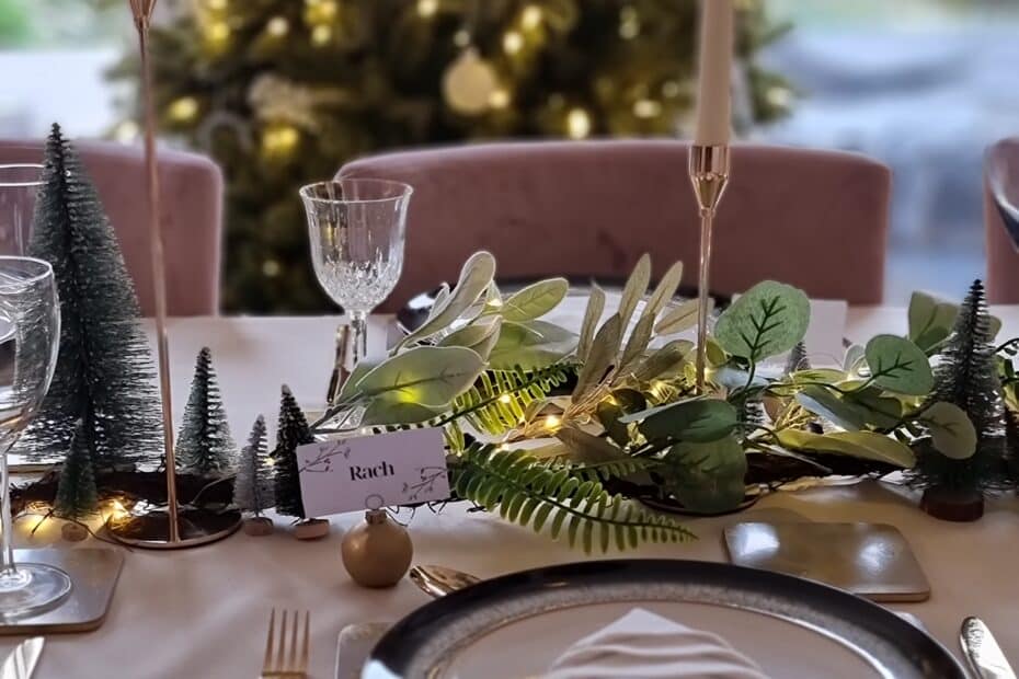 A festive dining table set with elegant plates, crystal glasses, and gold cutlery is perfect to Get Your Home Ready for the Holiday Season; twinkling fairy lights, mini pine trees, and a Christmas tree create a magical backdrop.