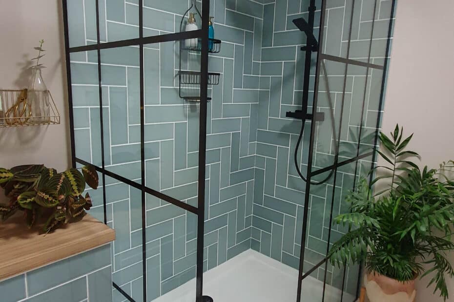 A modern bathroom with a glass shower enclosure featuring black frames, green geometric wall tiles, and stylish touches that add considerable value to your home, including potted plants, a speckled floor, and a wooden shelf with decor.