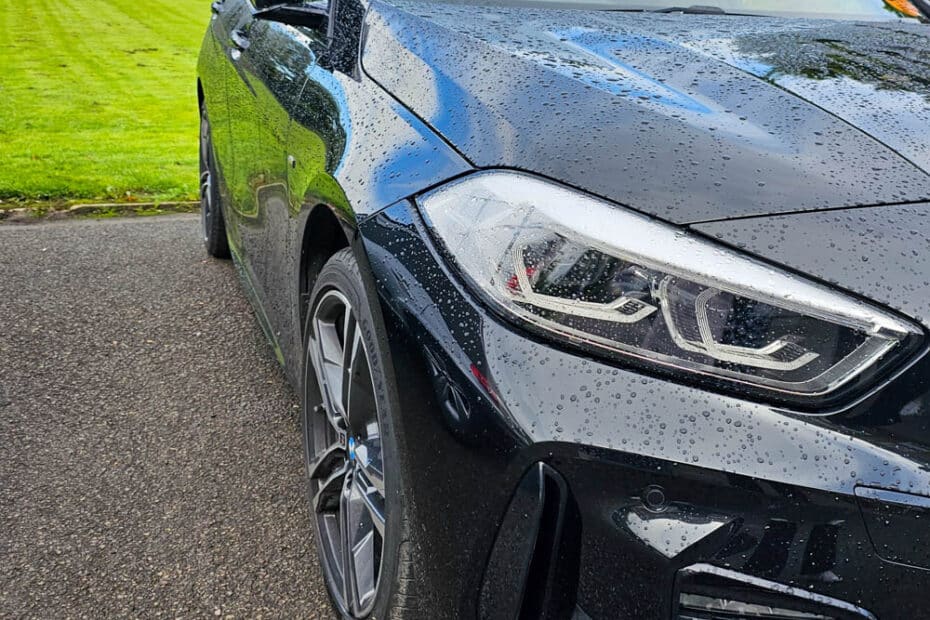 A shiny black car with rain droplets on its surface is parked on wet tarmac next to a green, well-manicured lawn and trees—perfect for anyone who’s Clueless About Cars but loves a striking scene.