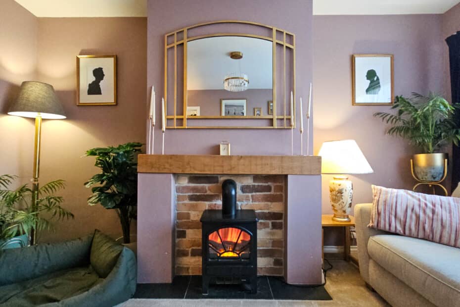 A cosy living room with a fireplace, mirror, and framed art above it. There are lamps, plants, a sofa with striped cushions, and a green pet bed on a beige carpet. The decor is warm and inviting.