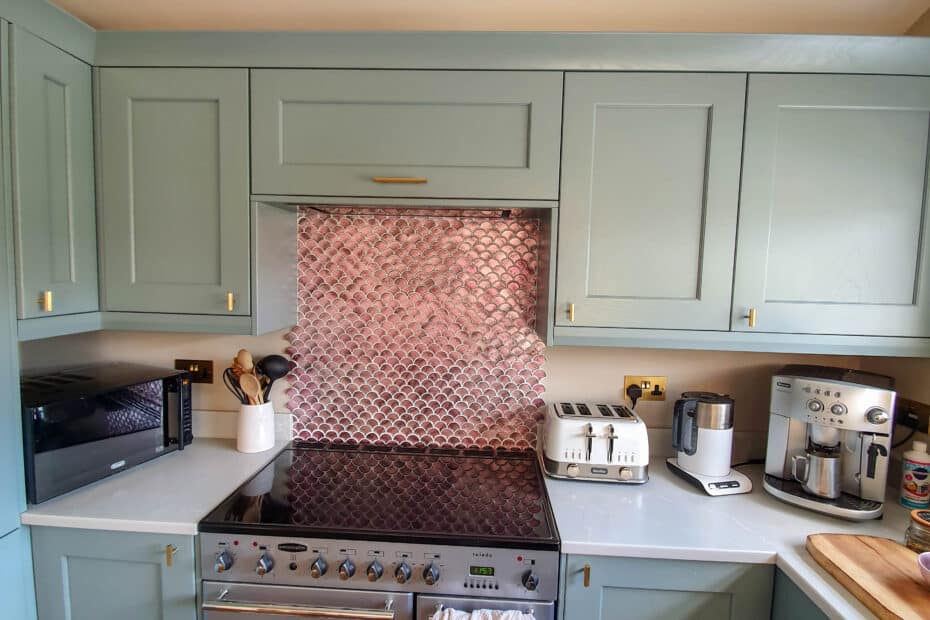 A modern kitchen with light green cupboards, a stainless steel cooker, a silver toaster, a coffee machine, a microwave, and utensils on white worktops, with a metallic splashback behind the cooker.