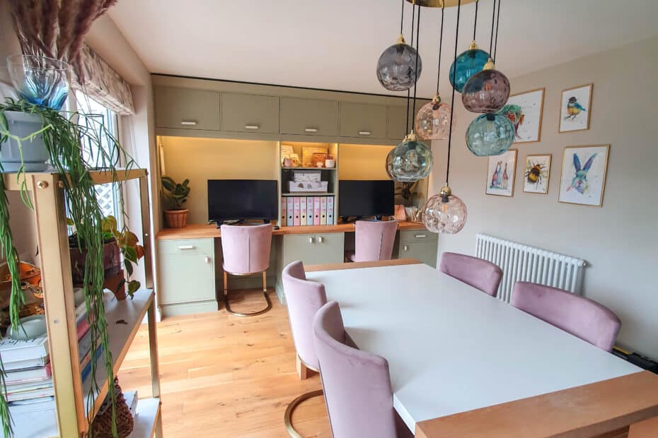 Modern home office with two desks, pastel green cupboards, and pink chairs around a white table. Hanging glass orb lights and shelves with plants make small rooms feel bigger, while large windows and wooden floors fill the space with natural light.