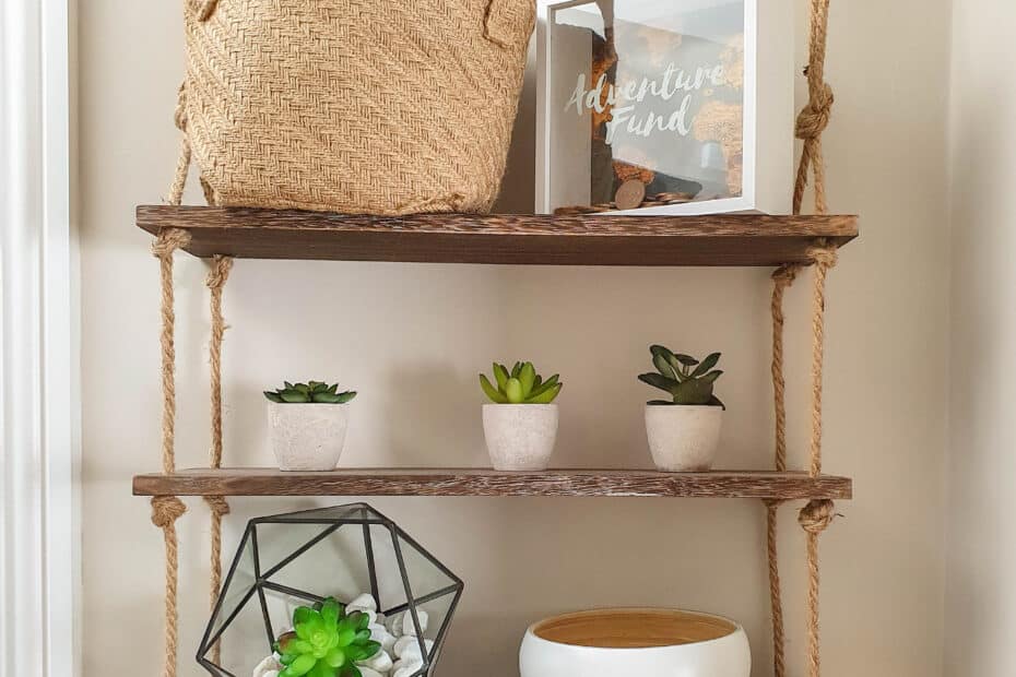 Three wooden shelves hang by ropes, displaying small potted plants, a geometric terrarium, a round bowl, and a woven basket—perfect decorative touches for anyone interested in affordable home building or budget home construction.