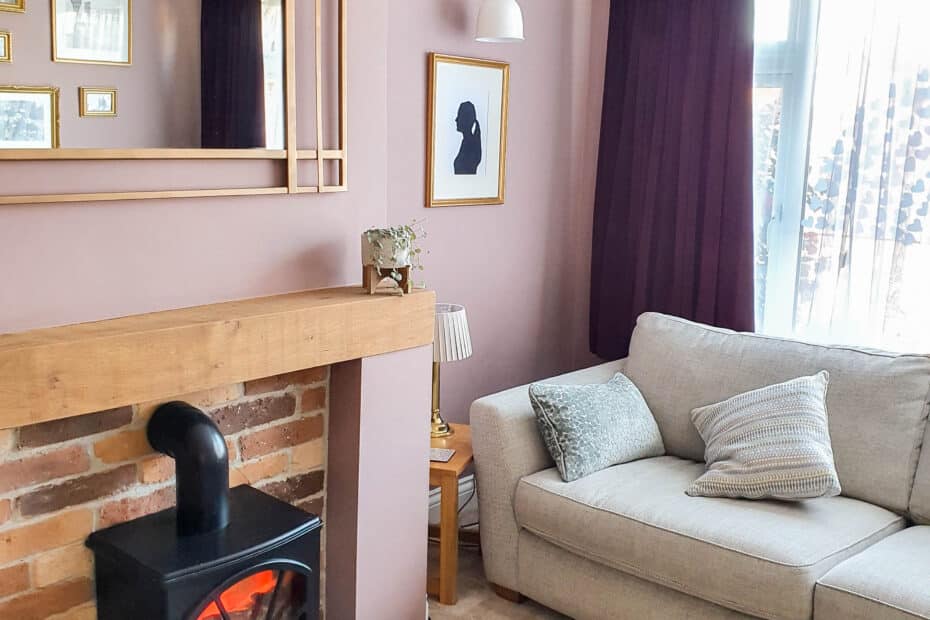 A cosy living room with a beige sofa, patterned cushions, a brick fireplace with a wooden mantelpiece, an electric stove heater, and clever seating layouts to make small rooms feel bigger, plus a large mirror and purple curtains over a sunlit window.