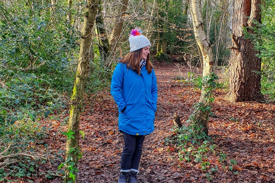 A person showcasing their personal style in a blue coat and grey hat stands on a leaf-covered path in a wintry forest, surrounded by bare trees and patches of green foliage.